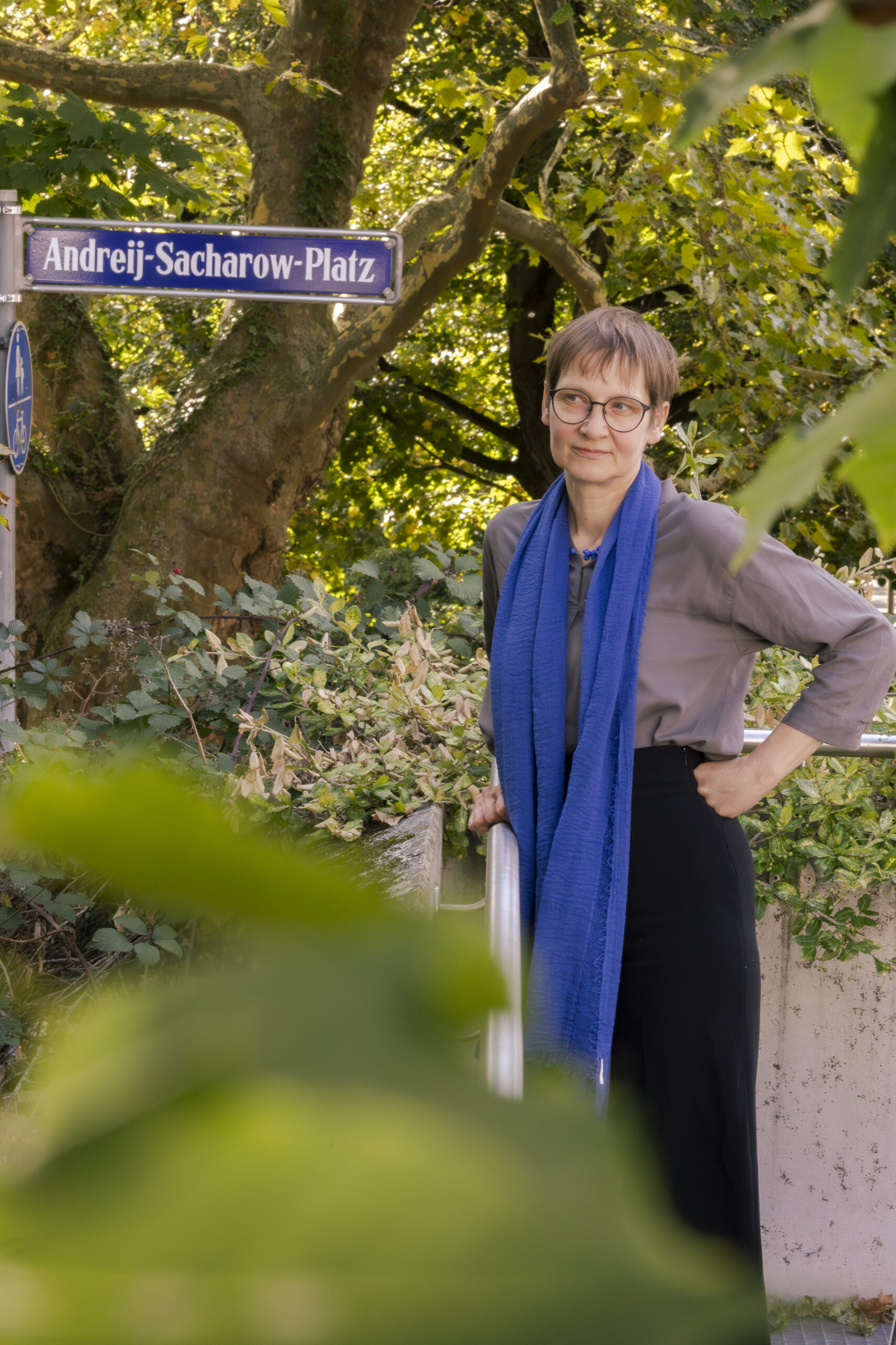 Prof. Dr. Eva Pils in frot of the Andreji-Sacharow-Platz sign surrounded by green.